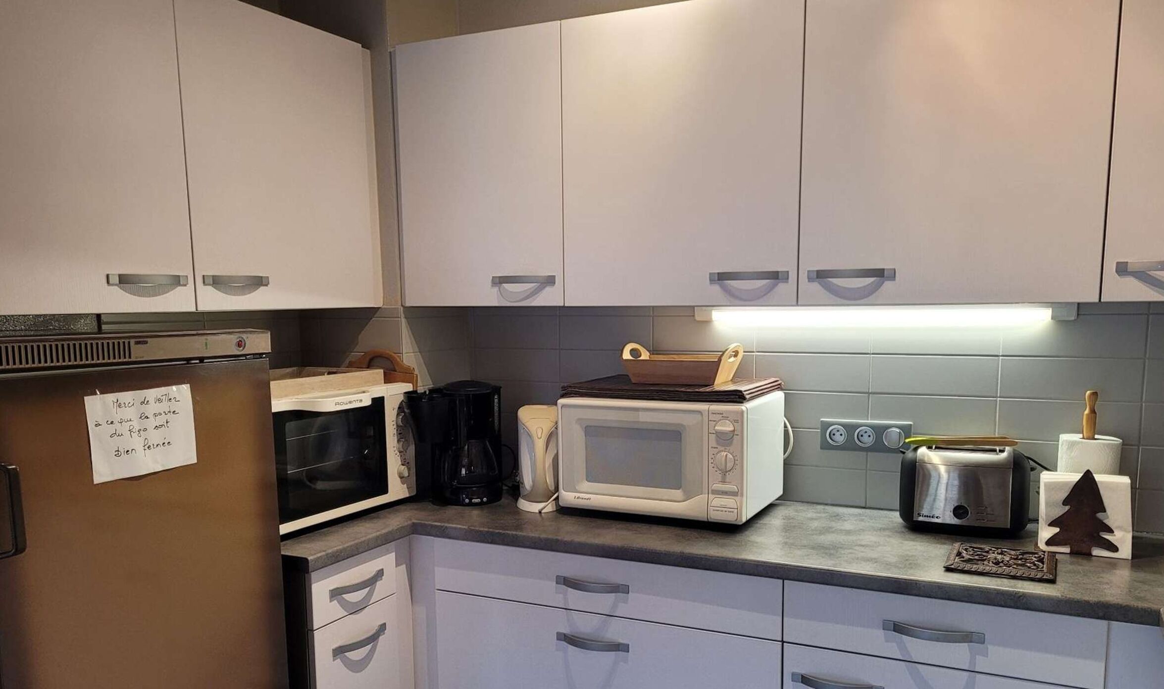 Eine Küche mit weißen Schränken, einer Mikrowelle, einem Toaster und einer Kaffeemaschine. Auf dem Kühlschrank liegt ein Zettel und auf der Arbeitsfläche ist eine kleine Weihnachtsdeko.