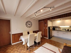 A dining area with a table and chairs, a clock on the wall, and a kitchen in the background. - Beautiful 2-room apartment in the heart of Le Grand-Bornand for 4 people (Le Grand-Bornand)