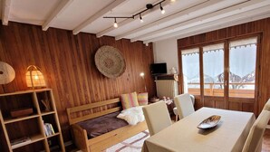 A cozy room with a wooden wall, a round woven wall hanging, a television, a dining table with chairs, and a view of snowy mountains through large windows.