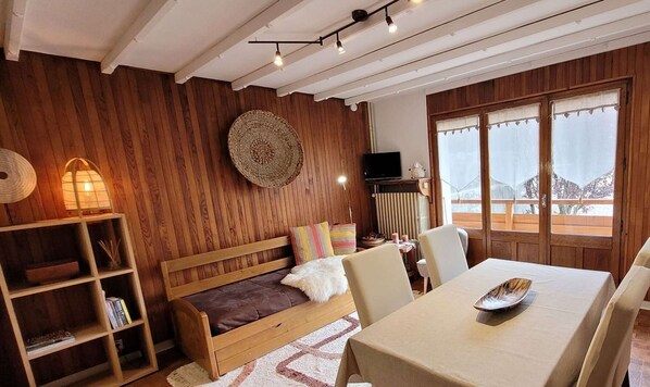 A cozy room with a wooden wall, a round woven wall hanging, a television, a dining table with chairs, and a view of snowy mountains through large windows. - Beautiful 2-room apartment in the heart of Le Grand-Bornand for 4 people (Le Grand-Bornand)