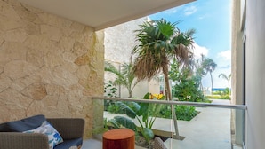 A balcony with wicker furniture, a round wooden stool, and a view of a tropical garden with palm trees. - Chic Beachfront Condo Ocean View (Akumal)