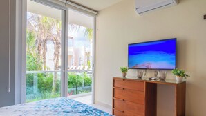 A bedroom with a bed, a TV on a wooden dresser, a window with a view of palm trees, and an air conditioning unit on the wall. - Chic Beachfront Condo Ocean View (Akumal)