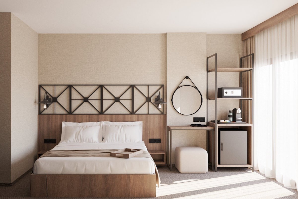 A hotel room with a large bed, a wooden headboard, a bedside table with a lamp, a small desk with a mirror, and a wardrobe.