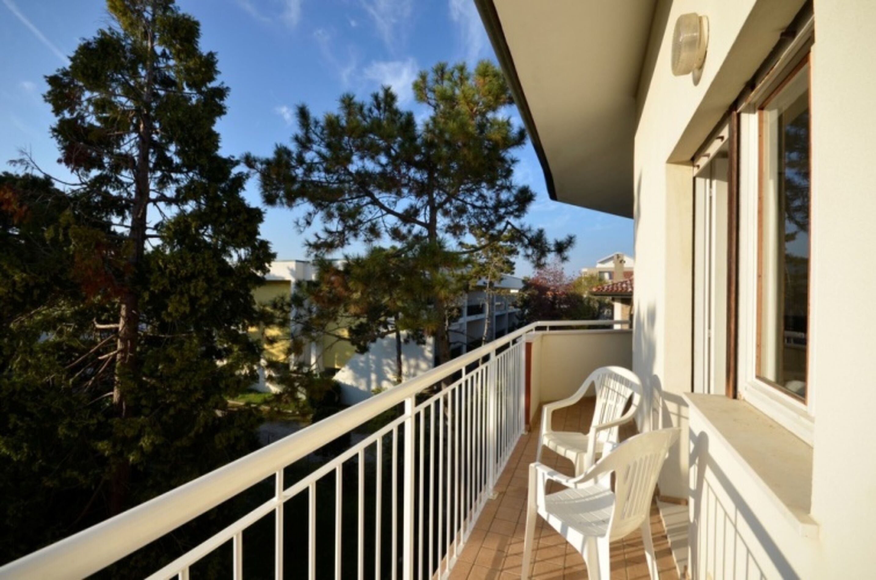 Un balcon avec des balustrades blanches, deux chaises en plastique blanches et une vue sur les arbres et les immeubles.