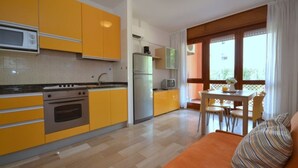 Cocina compacta con gabinetes amarillos, microondas, estufa y refrigerador. Cuenta con una zona de comedor con mesa y sillas, además de una ventana con cortinas.