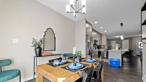 A modern dining area with a wooden table set for four, a kitchen in the background, and a staircase leading to another level. - 6 BR | Sleep 12 | Stadium & Rogers | Yard | Garage (Edmonton)