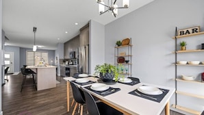 A modern kitchen with a central island, stainless steel appliances, and a dining area with a long table set for six. - Modern 4BR | Yard | Garage | Near Stadium & Rogers (Edmonton)