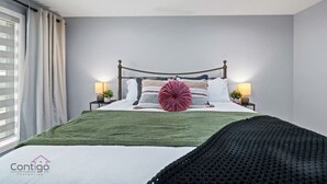 A bedroom with a bed, two bedside tables, a window with curtains, and a nightstand with a lamp. - Modern 4BR | Yard | Garage | Near Stadium & Rogers (Edmonton)