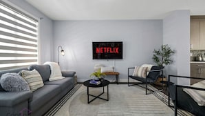 A modern living room with a grey sofa, a black coffee table, and a TV displaying 'NETFLIX'. - Modern 4BR | Yard | Garage | Near Stadium & Rogers (Edmonton)