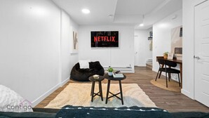A modern living room with a black bean bag, a small round table with books, a TV displaying 'NETFLIX', and a staircase leading to another room. - Cozy 2BR Bsmt Suite | Near Commonwealth & Rogers! (Edmonton)