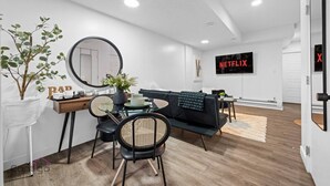 A modern living room with a dining area, a black sofa, a round mirror, and a wall-mounted TV displaying 'NETFLIX'. - Cozy 2BR Bsmt Suite | Near Commonwealth & Rogers! (Edmonton)