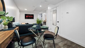 A modern, well-lit room with a glass-top dining table, black chairs, a wooden desk, and a potted plant. A large TV screen displays the Netflix logo. - Cozy 2BR Bsmt Suite | Near Commonwealth & Rogers! (Edmonton)