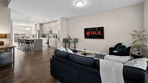 A modern living room with a dark sofa, a coffee table, and a TV mounted on the wall displaying 'NETFLIX'. - 4 BR | Yard | Garage | Near Commonwealth & Rogers! (Edmonton)