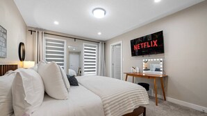 A hotel room with a bed, a desk with a mirror, and a wall-mounted TV displaying the Netflix logo. - 4 BR | Yard | Garage | Near Commonwealth & Rogers! (Edmonton)