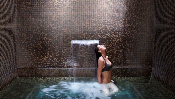 A person in a hot tub with a waterfall feature, surrounded by mosaic-tiled walls.