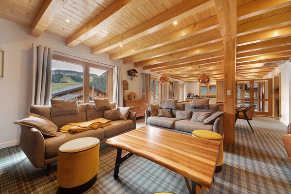 Amplia sala de estar con vigas de madera, un conjunto de sofás, una mesa de centro y vistas a un paisaje montañoso a través de grandes ventanales.