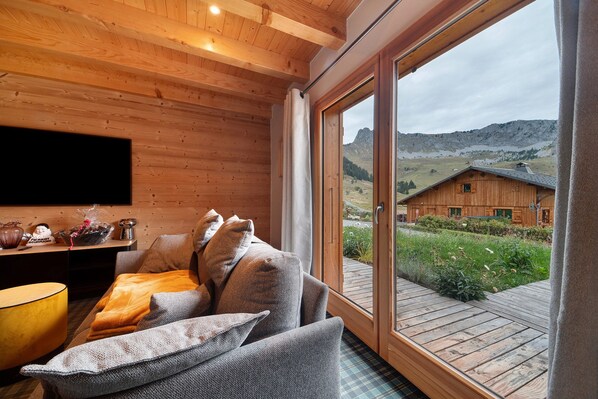 A cozy room with a wooden ceiling, a sofa with cushions, a small table with decorative items, and a large window offering a view of a wooden building and landscape.