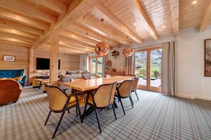 Amplia sala de estar con techo de madera, una mesa de comedor con sillas y vistas a través de grandes puertas de cristal.