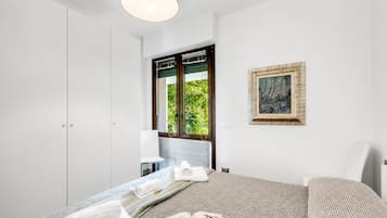 Ett hotellrum med en säng, en stol, ett fönster med persienner, en tavla och en taklampa.