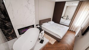 A modern hotel room with a large oval bathtub, a bed with white linens, a wooden floor, and a marble wall. - Ideal Apartments (Banja Luka)