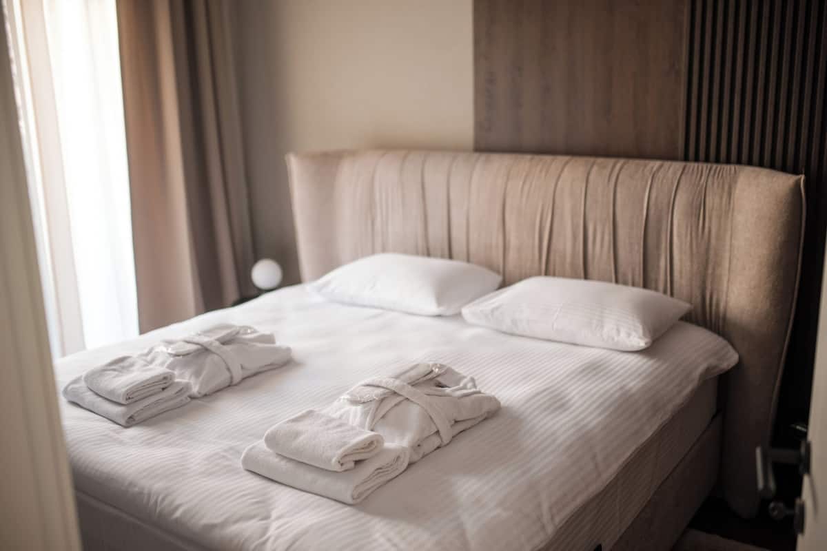 Een net opgemaakt bed met witte lakens en handdoeken, een houten hoofdbord en een raam met gordijnen.