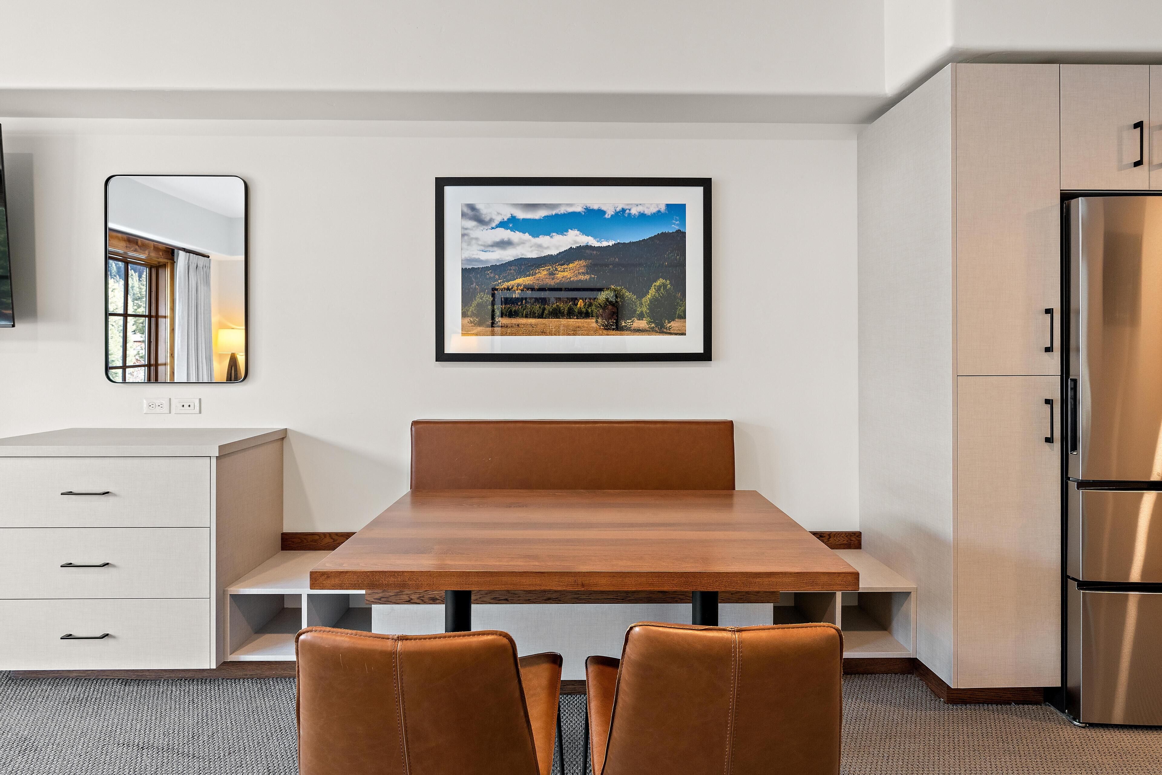 A modern kitchen with a wooden dining table, two chairs, a refrigerator, and a framed landscape picture on the wall.
