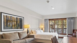 Habitación de hotel con cama, sofá, mesa de comedor y un amplio ventanal con cortinas.