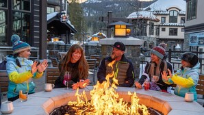 A group of people around a fire pit in an outdoor setting with buildings in the background. - Lodge Studio | Exclusive Tamarack Perks (Tamarack)