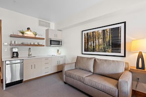 A modern kitchen with a built-in microwave, a coffee machine, and a sink. There is a sofa and a framed picture of a forest on the wall.