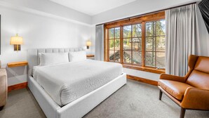 A bedroom with a large bed, a wooden headboard, a window with curtains, a brown leather chair, and two bedside lamps. - Moonlit Oak | Includes Two 1-Day Lift Tickets (Tamarack)