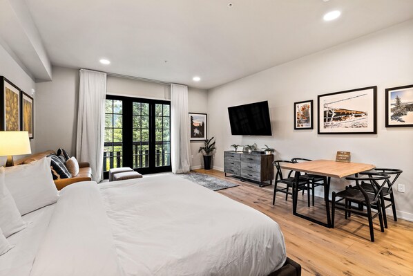 A modern bedroom with a large bed, a dining area with a table and chairs, and a TV mounted on the wall.
