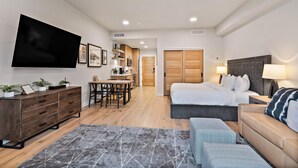 Habitación de hotel moderna con televisión de pantalla plana, cama con cabecera gris, sofá, mesa de comedor con sillas y zona de cocina.