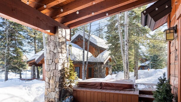 A wooden deck with a stone pillar, a bench, and a view of a cabin in a snowy forest. - Four Reasons | Exclusive Tamarack Perks (Tamarack)