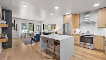 A modern kitchen with a central island, wooden cabinets, stainless steel appliances, and a dining area with a view of trees.