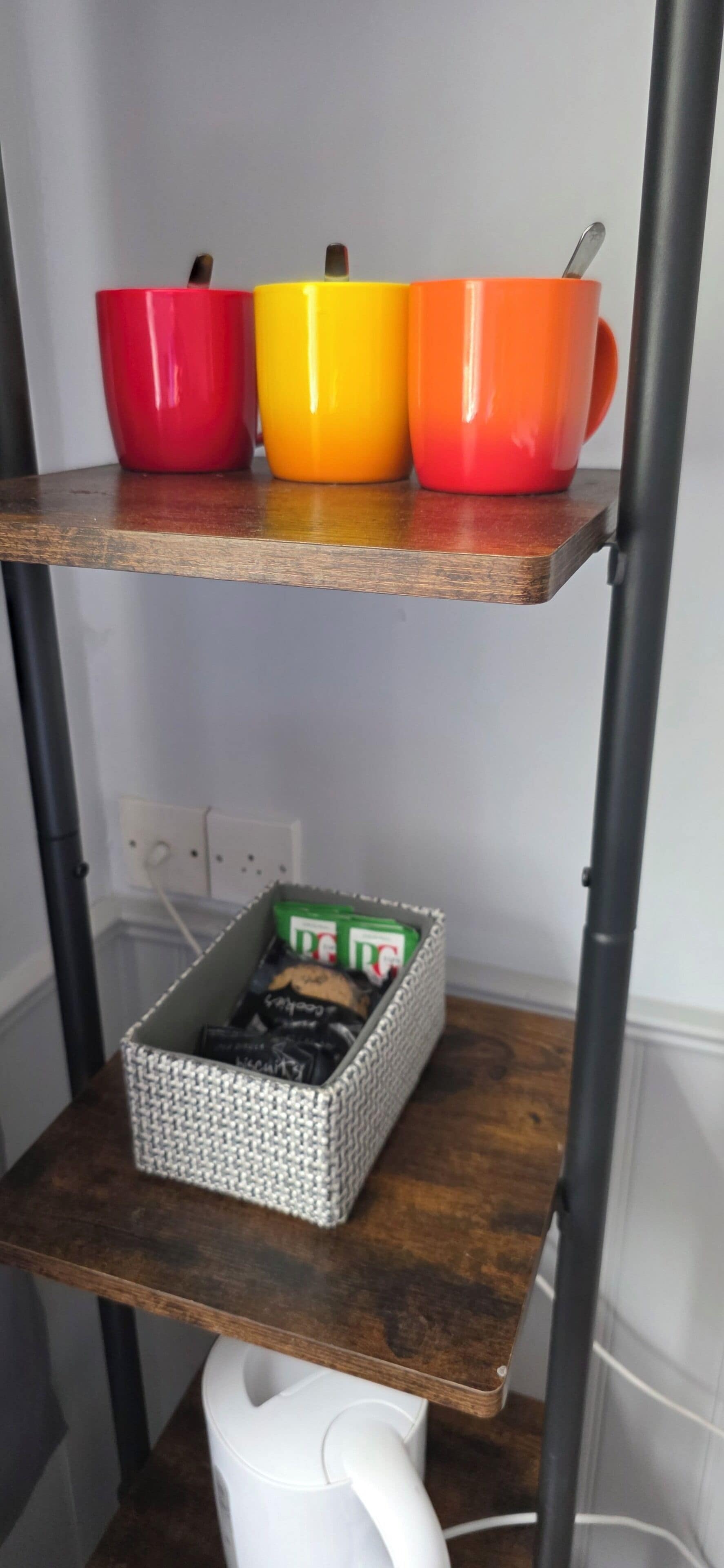 A shelf with three colorful mugs and a basket of snacks.