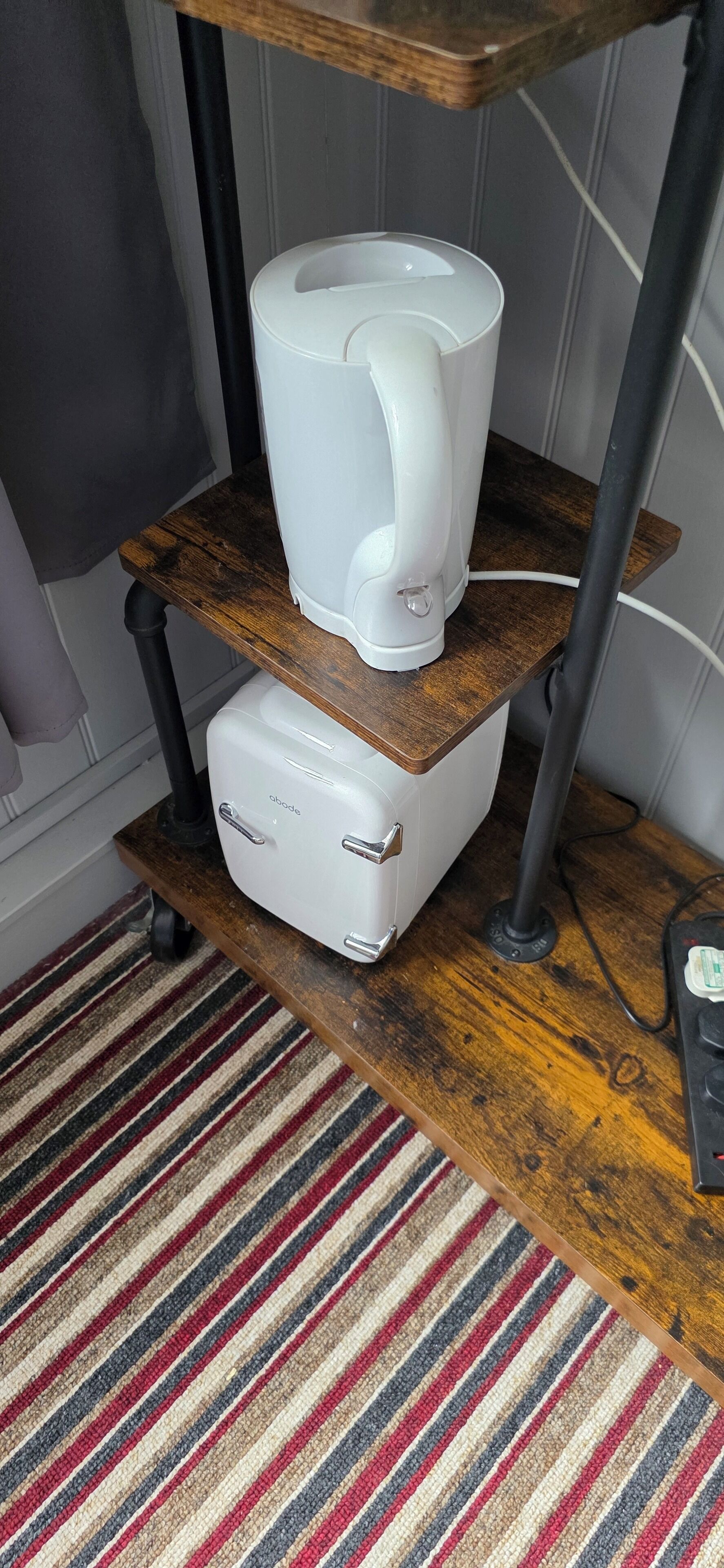 Un área de cocina con una cafetera sobre un estante de madera, un electrodoméstico blanco debajo y un tapete a rayas en el suelo.