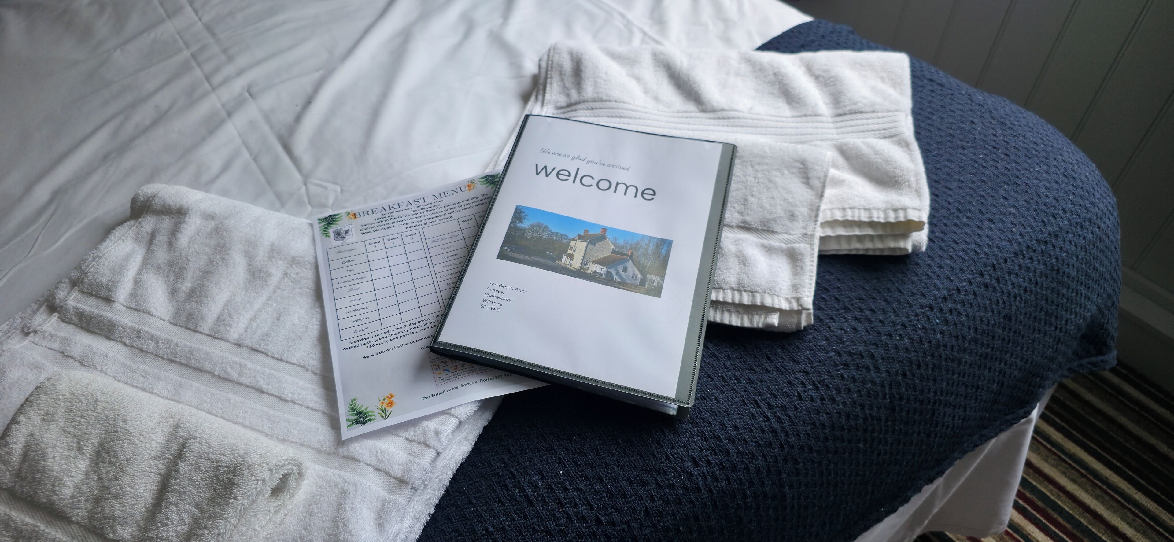 Ein ordentlich bezogenes Bett mit einem Willkommensbuch, einer Speisekarte für das Frühstück und einem gefalteten Handtuch.