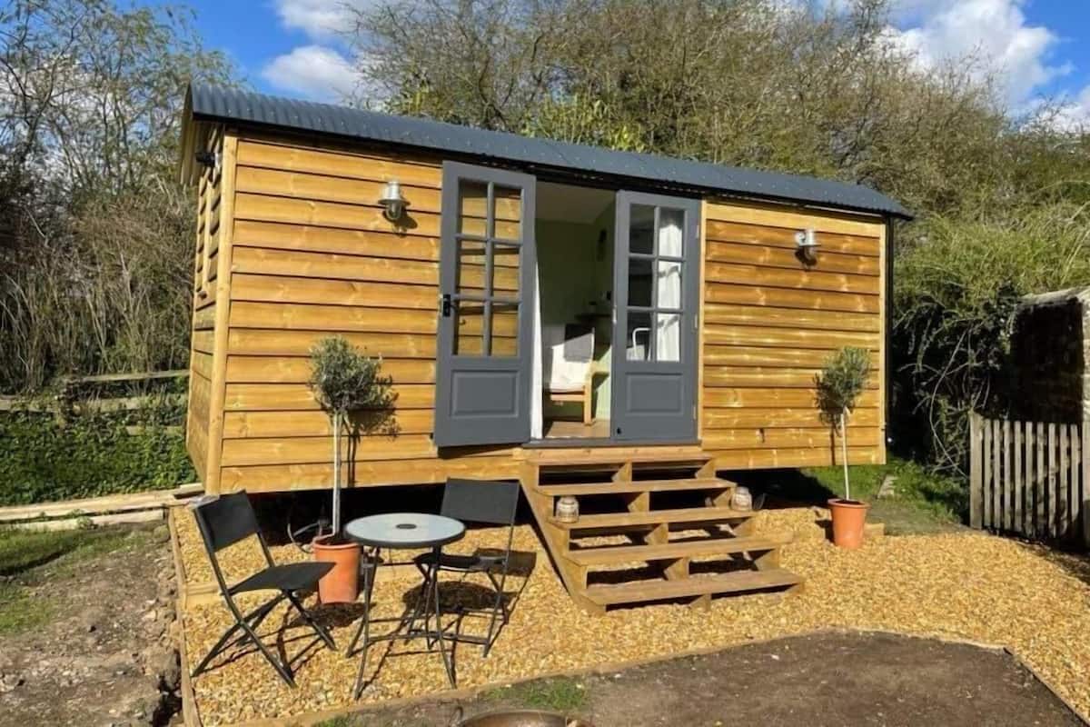 Une cabane en bois avec une porte coulissante, un petit patio avec une table et des chaises, et deux plantes en pot.