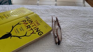 Ein gelbes Buch mit dem Titel "The Hunting Party" auf einem weißen, gesteppten Tisch, neben ihm eine Brille. Blick auf einen Balkon mit weißen Stühlen und Meerblick.
