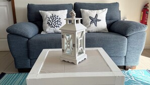Ein Wohnzimmer mit grauem Sofa, weißen Kissen, einem weißen Tisch mit Laterne und einem blauen Teppich.