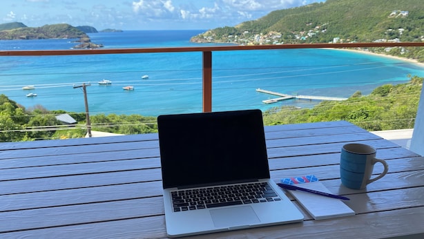 Ein Laptop auf einem Holztisch, im Hintergrund ist ein Strand und Boote zu sehen.