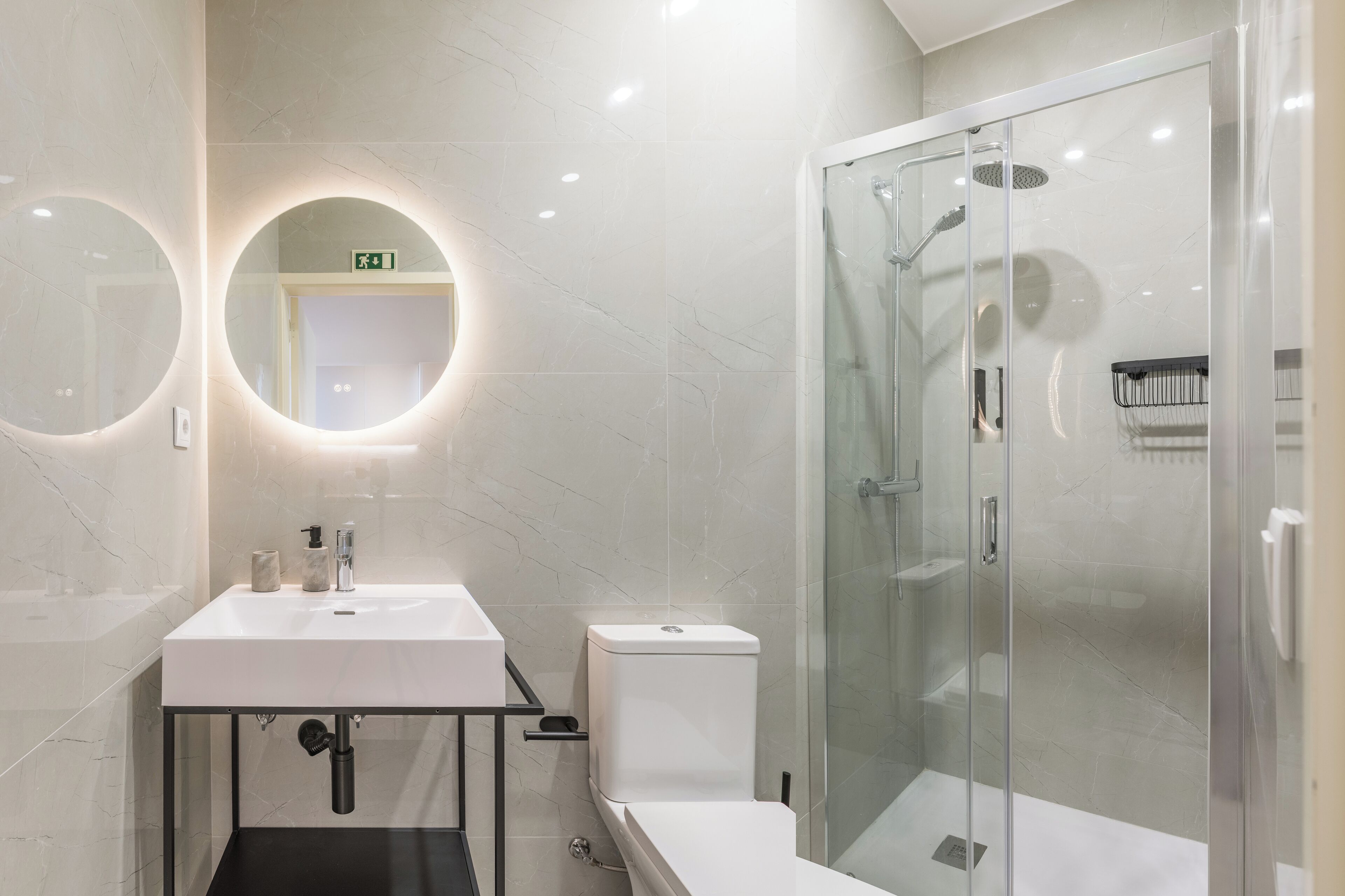A modern bathroom with a glass-enclosed shower, a white sink with a marble countertop, and a round mirror with a circular light.