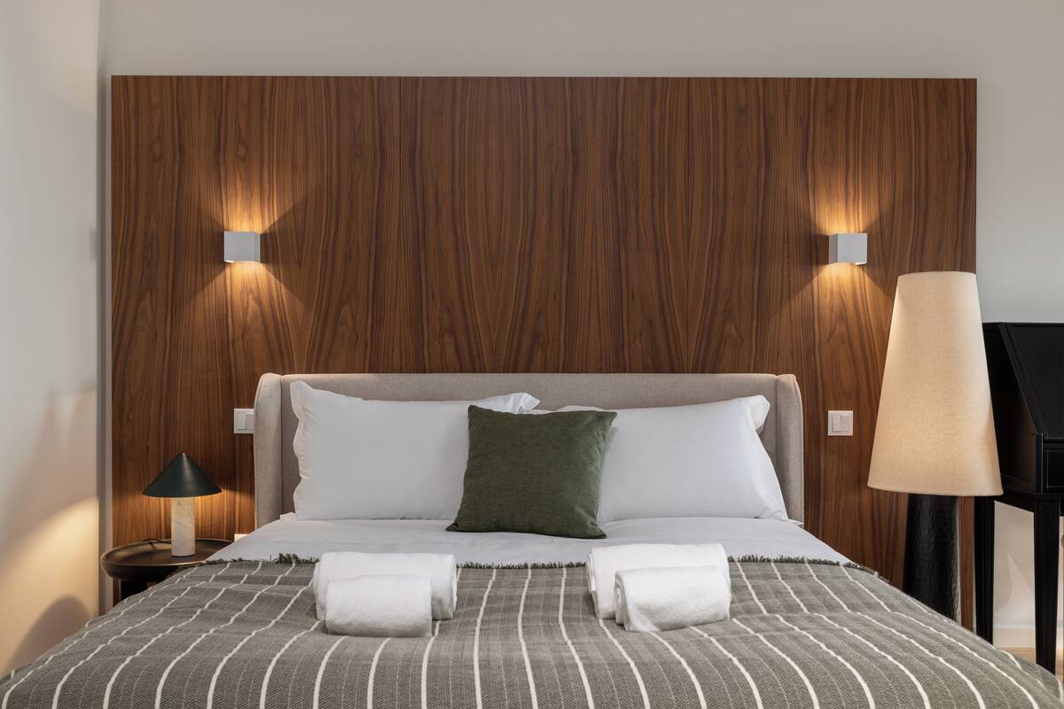 Una cama bien hecha con una manta a rayas, dos almohadas y dos toallas enrolladas.
