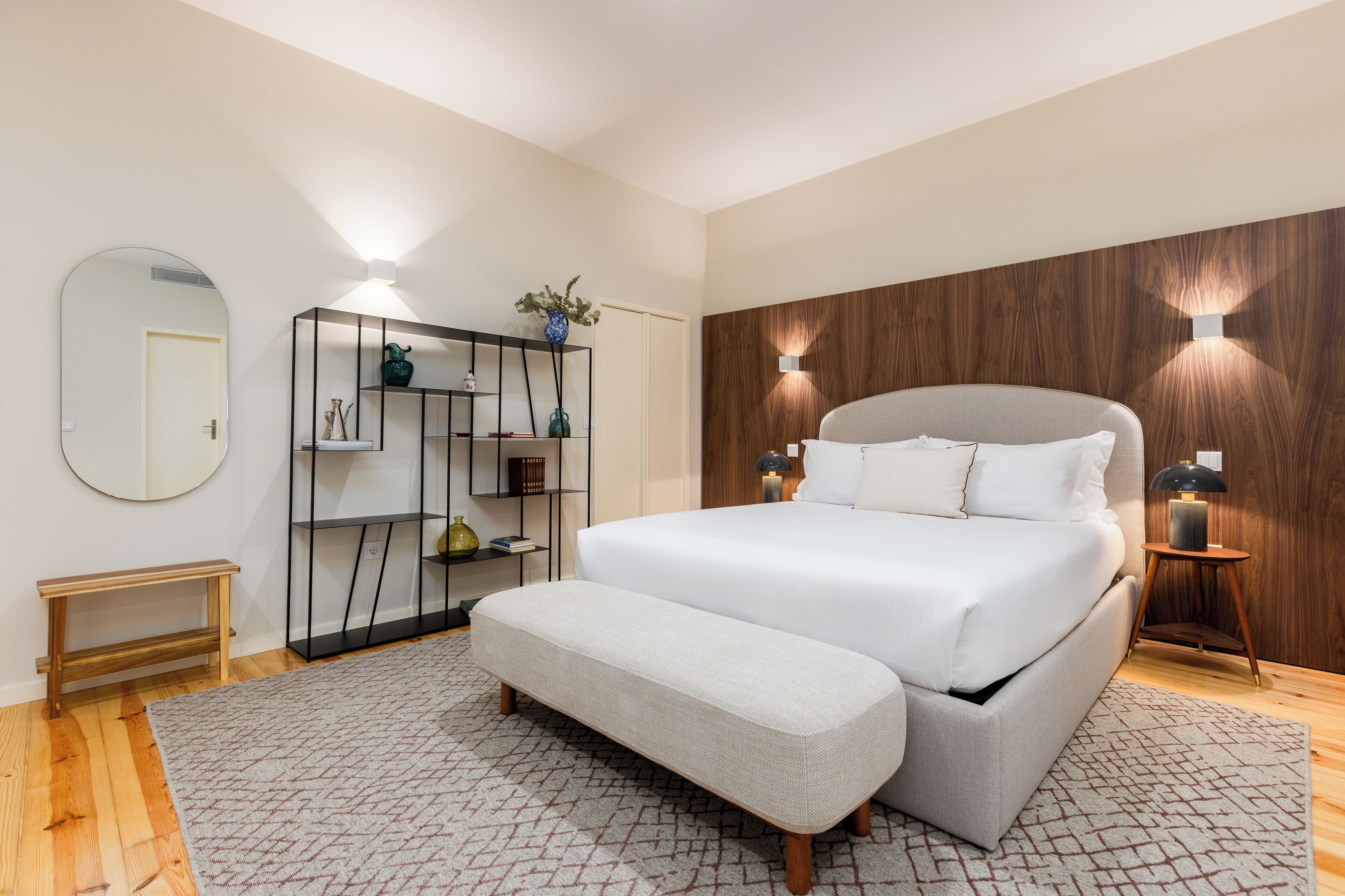 Une chambre moderne avec un grand lit, un banc, un miroir, une armoire et des lampes fixées au mur.