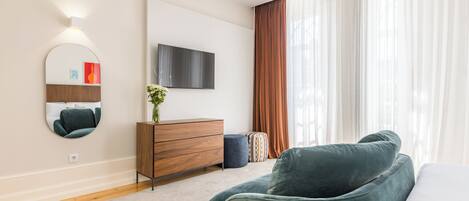 A modern bedroom with a large bed, a TV mounted on the wall, a wooden dresser, and a large window with curtains.