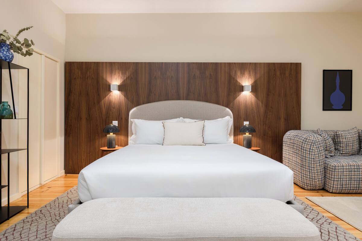 Un dormitorio moderno con una cama grande, dos lámparas de noche, un cabecero de madera y un sofá a cuadros.