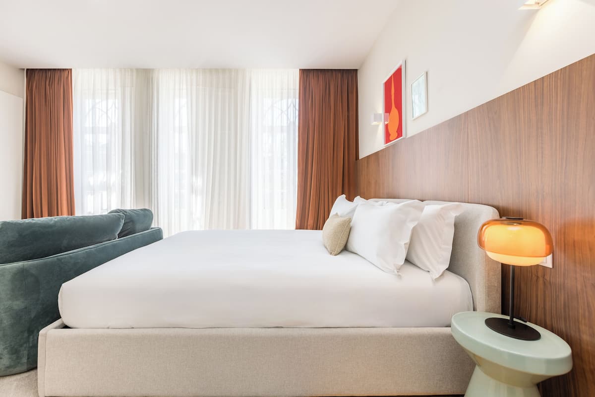 Habitación de hotel con una cama grande, una mesita de noche con lámpara, un sofá y vistas al exterior a través de cortinas traslúcidas.
