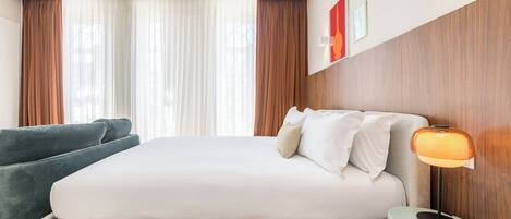 A hotel room with a large bed, a bedside table with a lamp, a sofa, and a view of the outdoors through sheer curtains.