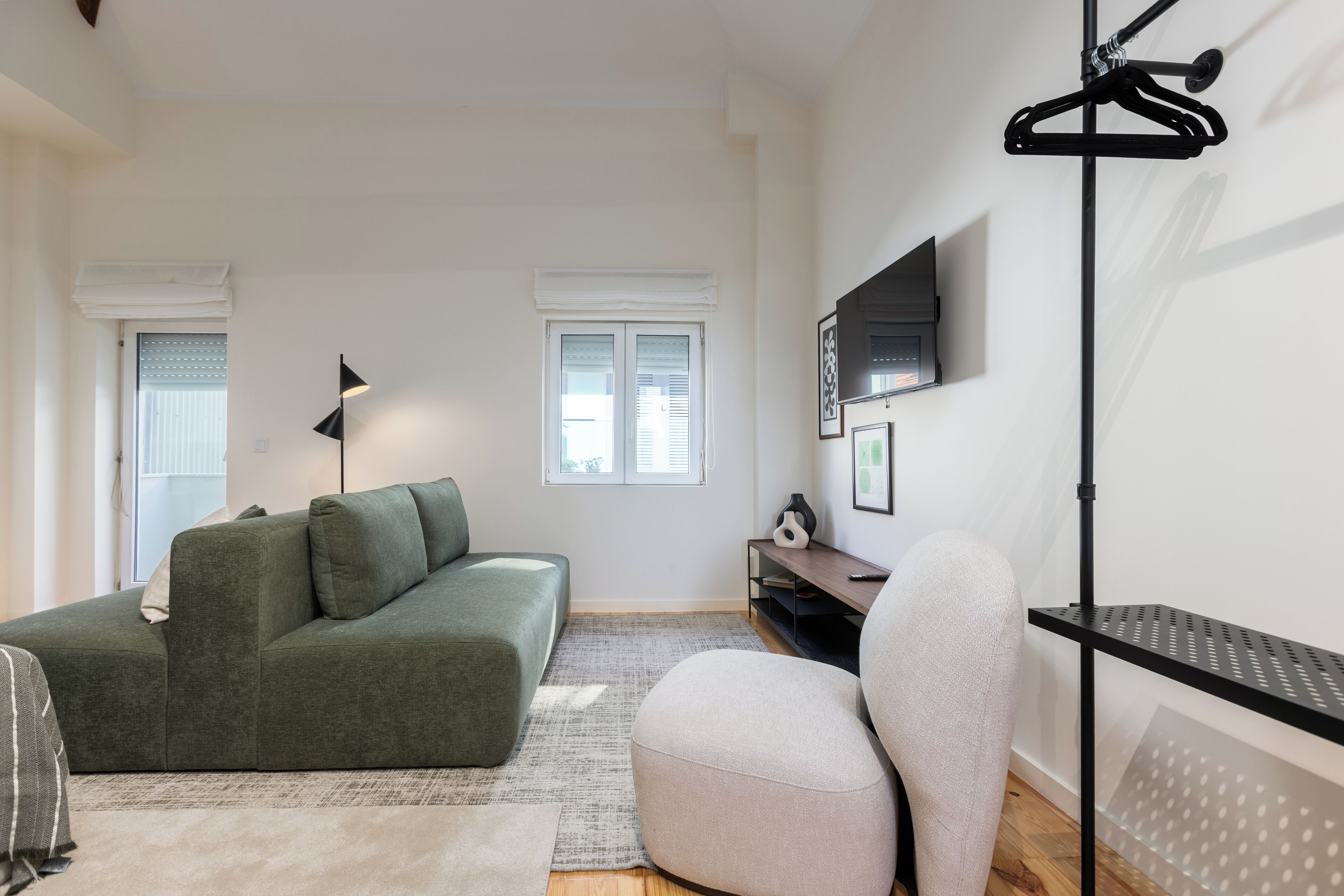 A modern living room with a green sofa, a light-colored armchair, a black floor lamp, and a TV mounted on the wall.