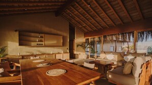 Un vaste espace de vie comprenant une grande table en bois, des sièges confortables, un téléviseur fixé au mur et une vue sur l’extérieur grâce à de grandes fenêtres.